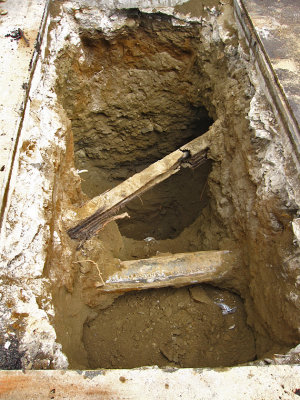 Hole is dug in the middle of the street, exposing phone line boxed in wood, <br>and water(?) pipe