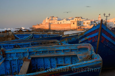 Blue fishing boats and Atlantic Ocean surf with the ramparts of Essaouira Morocco at sunset