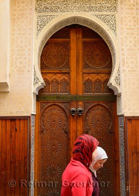 Two women in red hijab passing ornate wooden doors of a Mosque in Fes Medina Morocco
