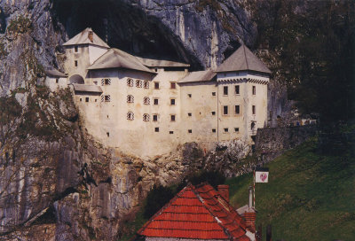 Photo showing the same view as the postcard. The village in front of the castle is named Predjama.