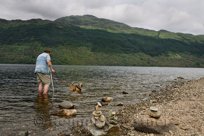 Convalescing man walking with crutch and dog on leash in Loch Lommond freshwater lake at Ben Lamond mountain Scotland UK