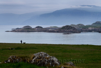 Woman walking her dog with view of Isle of Mull cliffs and Ben More mountain over Sound of Iona from Isle of Iona Abbey at dusk 