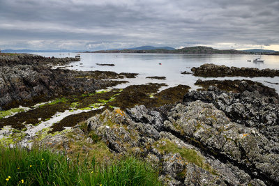 Sand beach and rocky shore under clouds on Isle of Iona with boats on Sound of Iona and Fionnphort Isle of Mull mountains Scotla