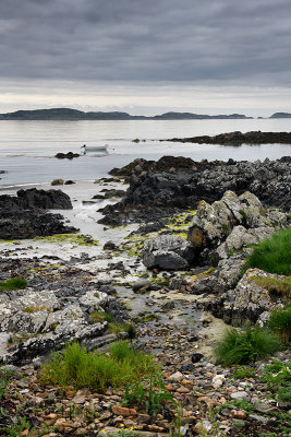 Sand beach and rocky shore and clouds on Isle of Iona with boat on Sound of Iona past Isle of Mull in Inner Hebrides Scotland UK