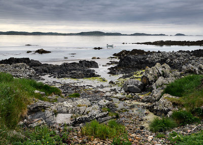 Sand beach and rocky shore and clouds on Isle of Iona with boat on Sound of Iona Inner Hebrides Scotland UK