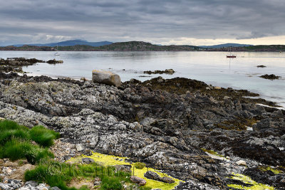 Rocky shore of Isle of Iona with sailboats on Sound of Iona and view of Fionnphort Isle of Mull mountains Inner Hebrides Scotlan