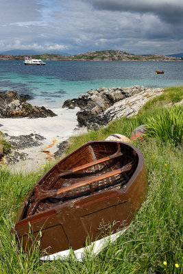 Sand beach and rocky shore of Isle of Iona with beached boat and view of Isle of Mull mountains of Ben More and tour boat on Sou