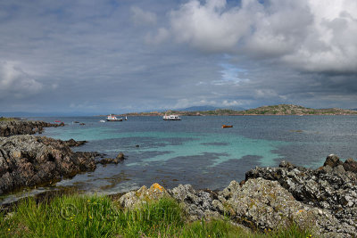 Rocky shore of Isle of Iona looking at Isle of Mull and mountains of Ben More and tour boats in Sound of Iona Inner Hebrides Sco
