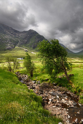 Stream to River Coupall valley with white cottage at foot of Buachaille Etive Mor mountains Glen Coe under dark clouds Scotland 
