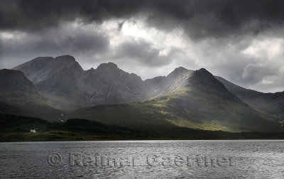 White house under Blaven mountains of Black Cuillin Hills with sun rays under dark clouds at Loch Slapin Isle of Skye Scotland U