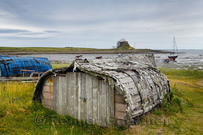 Weathered hull of shed made of overturned boat cut in half on Holy Island with Lindisfarner Castle ruins under renovation Englan