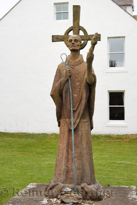 Statue of St Aidan first bishop of Lindisfarne monastery with white Manor House Hotel on Holy Island at the Priory ruins England
