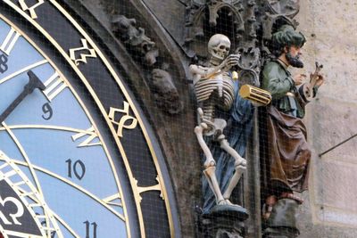 Death has an hourglass in one hand and rings a bell with the other one