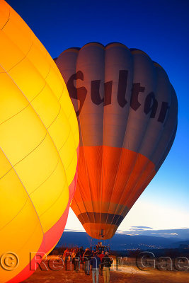 Tourists walking to an inflated hot air balloon while another is glowing from the heaters with Urgup Turkey city lights at dawn 