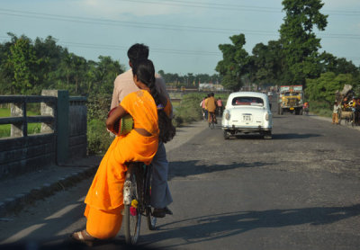 Woman in an orange sari riding sidesaddle on a bicycle, West Bengal