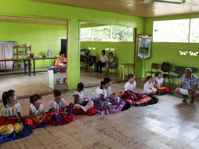 Students at a small, rural school. The teacher is wearing an orange blouse. The parents are sitting in the background.