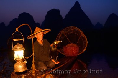 Fisherman holding cormorant on bamboo raft with lantern basket and net at dawn on the Li river Yangshuo China
