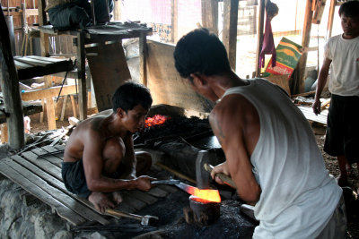 While one blacksmith holds the blade, the other pounds the edge of it.