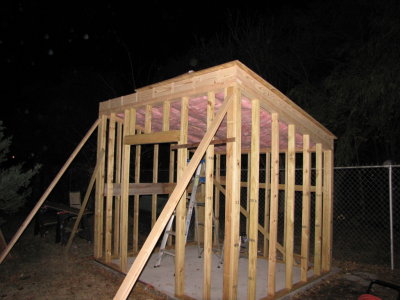 Once I got two pieces of plywood on the rafters, I moved them around and removed my temporary platform. I laid out the shed to use standard widths of plywood on the walls, but since the roof is angled and will have 2 edges to cover the siding and trim, the 4'x8' boards were not long enough and I had to add a 7 strip along the bottom before attaching the 4'x8' boards. I stapled in the insulation from the top, so it would hang down and fill the space between rafters. Then, I got the bottom strip and two full sheets of plywood up before it got too dark to see what I was doing.