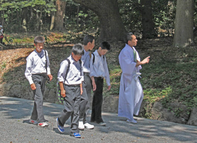Schoolboys and a monk approaching the Meiji Shrine inner complex on the gravel road entrance surrounded by cedars - Tokyo