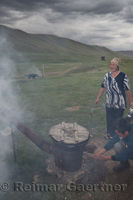Son stoking fire on outdoor oven stewing lamb with mother laughing on Assy Plateau Kazakhstan
