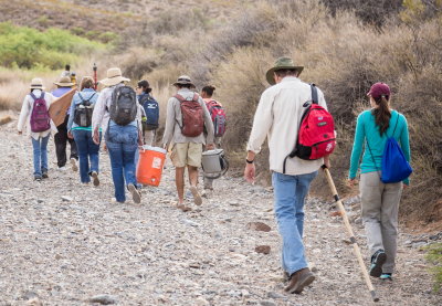 Crew carrying gear from vehicles to the site followed by visitors Dr. Warren DeBoer and Dr. Mary Alice Scott