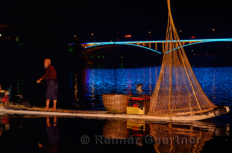 Cormorant fisherman on raft at night with lights of Yangshuo Bridge reflected in the Lijiang River China