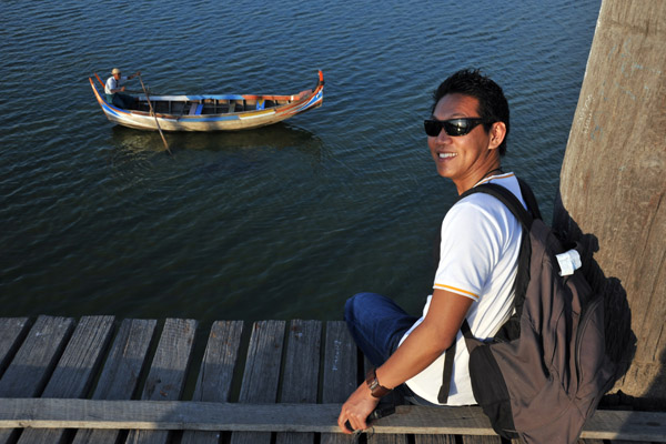 Dennis sitting on the edge of the Teak Bridge as a hopeful boatman waits patiently