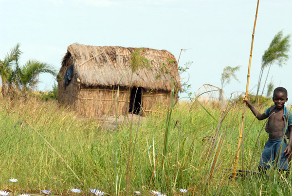 Boy with bamboo poles in front of their grass hut, Bangweulu Swamps
