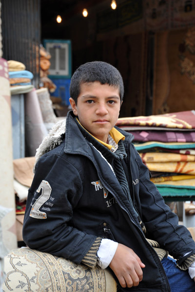 Kurdish boy leaning against rolled up carpets
