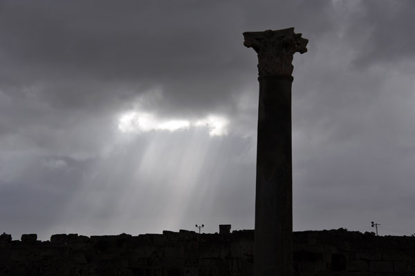 A ray of light shines through the dark clouds behind the silhouette of a lone column at Sabratha