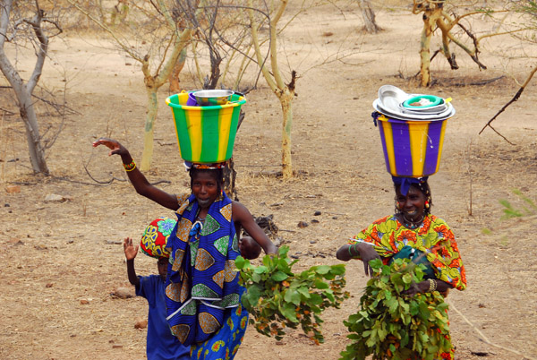A pair women in brightly colored clothing carrying buckets of dishes