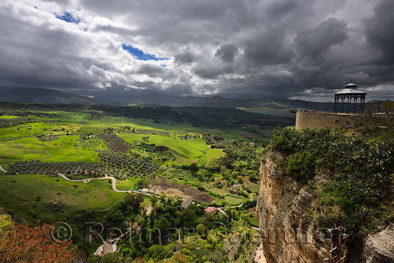 Dark clouds and dappled sunshine on green farm fields below lookout at  city of Ronda Spain