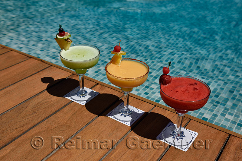 Three colorful alcoholic drinks on a deck next to a pool