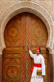Man in white robe and red cap and sash at door of Mosque with intricate stone carving and paint in Fes Morocco