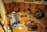 Woman working a weaving loom while others prepare the thread