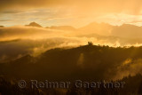 Golden clouds and fog at sunrise in the mountains of Kamnik Savinja Alps in Skofjelosko Hills with St Thomas church silhouette n