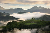 Rolling fog at sunrise in the Skofjelosko Hills with St Thomas church with mountains of Kamnik Savinja Alps near Ljubljana Slove
