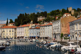 Pastel houses on Cankar Quay along the inner harbor of Piran Slovenia with moored boats and crenellated Town Walls on hilltop