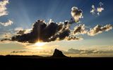 Rays of the setting sun light up the clouds and silhouette Shiprock