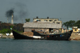 A pair of black wooden freighters in front of a belching smokestack on the outskirts of Dhaka