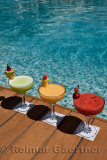 Red yellow and green cocktails on a wooden deck next to a pool