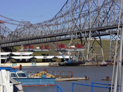 February 3rd, 2003 -- Here you can see that two deck sections have been lifted from the ship and are hanging from the bridge's suspension cables.  Protective covers, seen on the six sections yet to be unloaded, are removed prior to the lift.