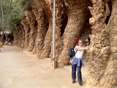 Judy holding up columns of stone that seem to be simulated palm trees - they form a cave-like passageway - in Parc Gell.